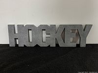 Three-dimensional decorative sign spelling 'HOCKEY' in uppercase letters with a textured gray finish, shown on a black surface.