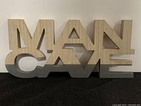 Photo of the front view of the 3D wooden 'MAN CAVE' sign showing the natural wood and gray painted letters on a black surface.