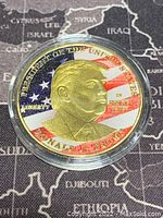 Front side of the gold colored medallion featuring a detailed embossed profile of former US President Donald Trump, American flag backdrop, and text.