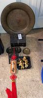 Lot contents grouped together on carpet including Asian decorative wall plates, cherub figurines, decorative ashtray, hanging wall art with red tassel and Chinese characters, and box with glass decanters and shot glasses.