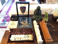 Chopsticks in wooden box on table alongside various Asian decorative items including ashtray, framed art, Buddha statuette and carved reliefs