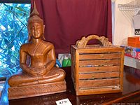 Photo of carved wooden Thai Buddha statue and carved wooden jewelry box with drawers on a dark wooden surface with maroon curtain background.