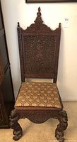 Front view of antique Burmese chair showing ornate carved wooden back with a carved figure and finial at top, cushioned seat with patterned upholstery.