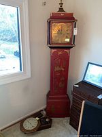 Full view showing red grandfather style floor clock standing against wall with smaller wall clock components on carpet