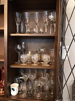 Wooden cabinet shelf with multiple types of glassware including stemware, snifters, and a ceramic mug visible on bottom shelf