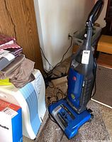 Blue Hoover upright vacuum cleaner shown standing on carpet with visible handle, controls, and upholstery.