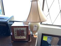 Photo shows a wooden mantel clock and a cream ceramic table lamp on a dark wood surface in front of a window.