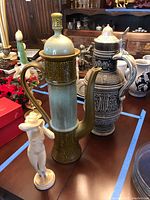 Photo showing three main items: ceramic coffee pot with green/beige glaze, West Germany beer stein with intricate black/white relief and metal lid, and white female figurine on round base.