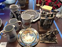 Wide view showing numerous ceramic items and bronze statues arranged on table, focus on mugs, pitchers, decorative plate, and figurines.
