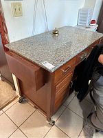 Front and side view of wooden rolling kitchen island with granite-like stone top, drawers, cabinet, and towel rack, showing condition and design details.