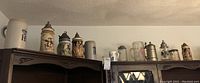 Wide view of 10 various beer steins and mugs including ceramic and glass types placed on top of a cabinet.