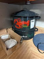 Two-piece black speckled metal stock pot with lobster and clam broth graphics and lids on shelf, white ceramic dishes in cardboard box, round blue speckled trivet on side.