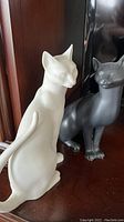 Two bisque cat statues, one white and one black, displayed on a wooden surface near dark furniture and a TV.