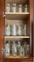 Three shelf cabinet containing several rows of clear glass Ball brand canning jars in various sizes and styles.