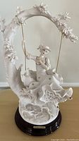 Full view of the Giuseppe Armani porcelain sculpture 'Young Lady on Swing' showing the young woman seated on ornate swing adorned with flowers.