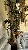 Full view of the faux wisteria tree showing cascading light pink flowers, green leaves, and beige planter near a window and couch.