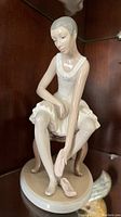 Front angled view of the Lladro ballerina figurine seated on a stool, showing her putting on ballet shoes.