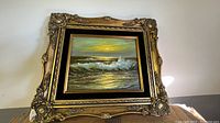 Frontal photo of oil seascape painting with ornate gold frame showing waves crashing and sunset sky.