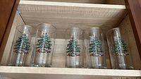 Five clear glass tumblers painted with a Christmas tree design by Spode, arranged in a row on a wood shelf.