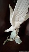 Side view of white turtle dove figurine perched with wings spread on a branch with large leaves.