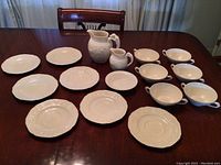 Photo showing entire lot of 14 pieces arranged on wooden table, including pitchers, bowls, saucers, and plates.