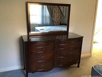 Full view of wooden dresser with a curved front, six drawers with brass handles, glass top, and large rectangular mirror mounted behind.