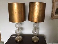 Pair of two table lamps with clear glass spiral orb bodies and gold metallic cylindrical lampshades on a wooden surface.