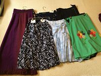 Six skirts hanging including green skirt with flower embroidery, blue plaid skirt, black skirt, black and white patterned skirt, dark purple pleated skirt, and purple skirt with vertical stripes and fringe.