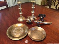 Full view of all seven yellow brass items on table including two candlesticks, plates, bird figurine, bell, and container.