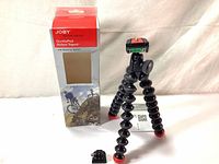 Joby GorillaPod Action Series tripod standing with its packaging and an accessory mount on a white cloth background.