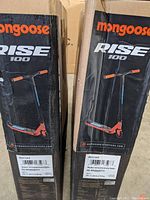 Two Mongoose Rise 100 scooters in original boxes standing upright showing front and top sides with branding and model name.