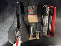 Top view of black and white carbon fiber bicycle saddle, carbon fiber cage still on backing card, Topeak AirStik 2-stage mini pump in box.