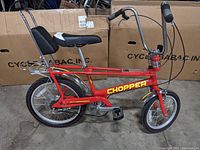 Side view of the Raleigh Chopper style bicycle showing red frame, yellow branding, black banana seat with backrest, rear luggage rack, and front small wheel.