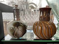 Two artisan ceramic Lampe Berger lamps on glass shelf: one brown with carved swirls and cutouts, one beige with raised leaf design and silver metal top.