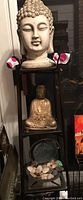 Photo showing three items stacked on a black shelf: Buddha head bust on top shelf, seated Buddha figurine on middle shelf, and round fountain basin with rocks on bottom shelf.
