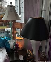 Photo of multiple lamps including ceramic lamp with white shade, painted metal lamp with black shade, and smaller decorative tulip shape lamp with hummingbird figurine base on table.