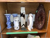 Wide view of shelf showing multiple ceramic and pottery items including the penguin figure, vases (blue and white pair, abstract brown textured), and a black and white patterned small vase with label.