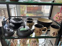 Shelf display showing multiple pottery items including bowls, mugs with leaf designs, plates, and a black cast iron teapot