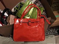 Red patent leather handbag, rectangular with buckle closure and a red tag hanging from the handle, surrounded by other bags in a cardboard box.