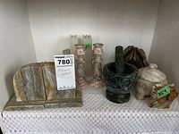 Wide view of the lot items together on a lace-covered shelf. Items include stone bookends, candle holders, mortar and pestle, alabaster jar, and turtle figure.