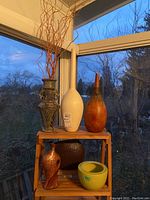 Six vases displayed on wooden shelves near a window at dusk, showing details on shape and texture including dried floral sticks in the cream vase.