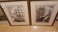 Photo of two framed monochrome art prints of sailing ships leaning against a wall.