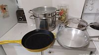 Yellow enameled cast iron grill pan with ridged interior, Lagostina stainless steel pasta pot with lid, aluminum frying pan, juice jug on kitchen counter