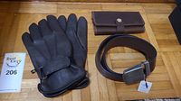 Auclair leather gloves, Post Card leather belt, and brown leather bifold wallet on wooden floor