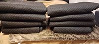 Ten black moving blankets stacked in two piles on a beige and floral textured surface. The blankets have a quilted zigzag stitch pattern, suitable for cushioning furniture during moves.