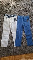 Two pairs of kids pants, one white with striped belt, one blue, both with tags, laid flat on carpet.