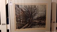 Full frontal view of framed and matted charcoal and pen drawing depicting a riverside cityscape with buildings, leafless trees, and bridge