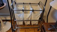 Full front view of the rolling laundry cart showing three large canvas laundry bags hanging from metal rods and the bottom wire shelf with caster wheels.