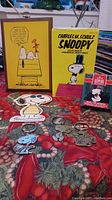 Full lot display includes yellow wooden wall hanging, yellow book box, Snoopy ornament in box, three metal keychains on table with holiday cloth backdrop.