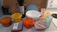 Overall photo of various Tupperware items including orange and yellow bowls with lids, a white bowl with handle and lid, pastel colored glasses, pink storage containers, and iced coffee maker
